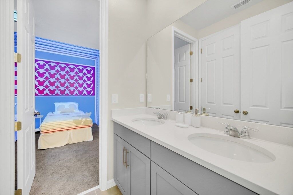 A bathroom with two sinks, a large mirror, and a door leading to a bedroom with a bed and a blue wall with a pattern.