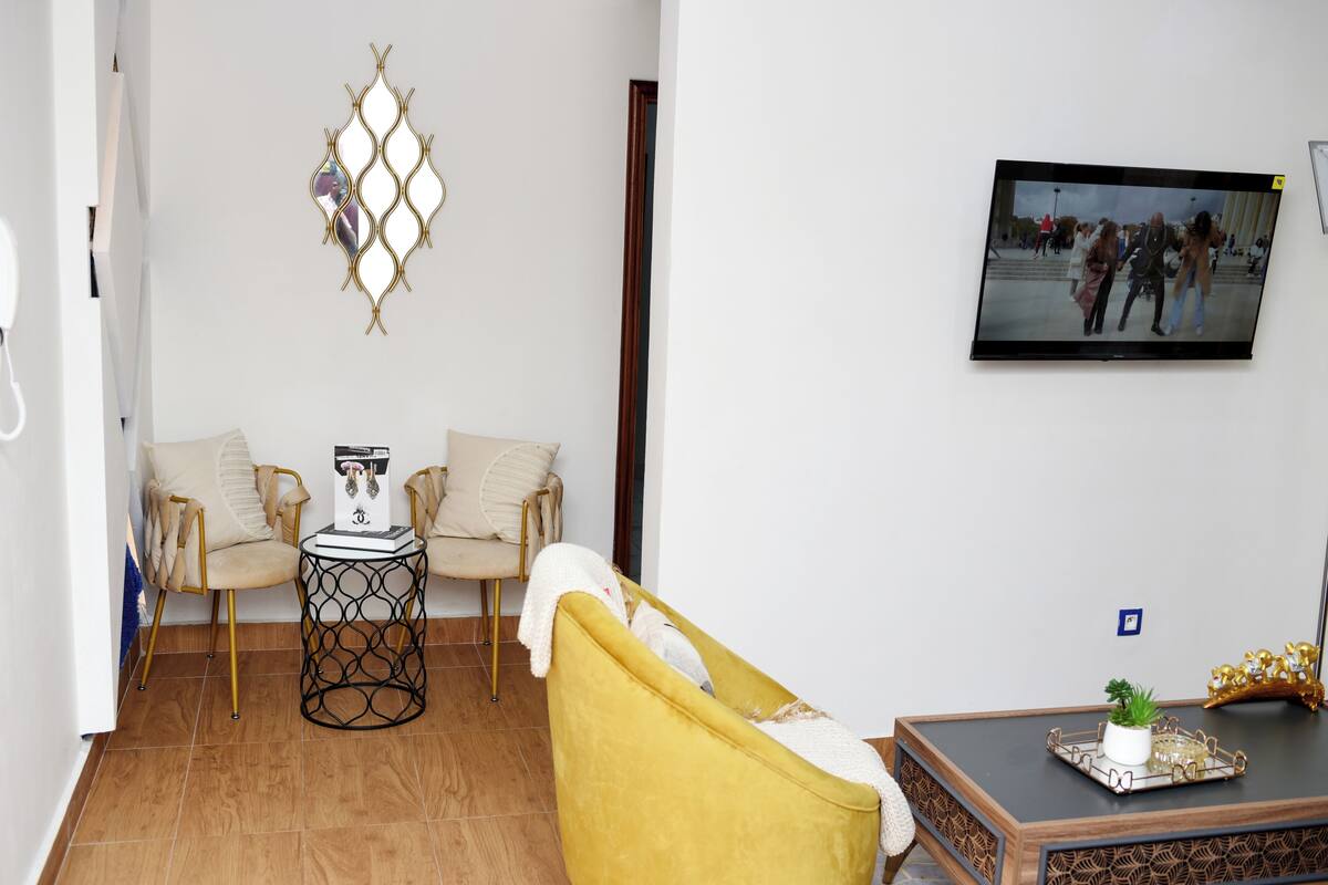 A living room with a yellow armchair, two beige armchairs, a small round table, and a television mounted on the wall.