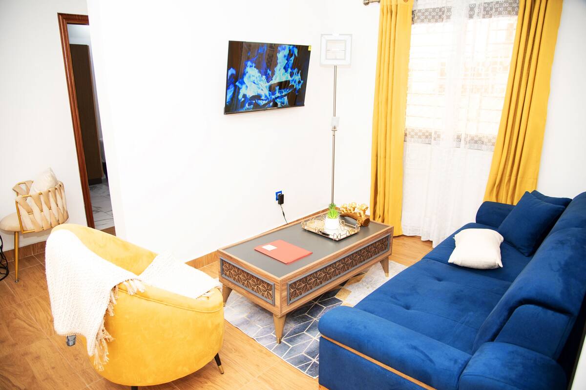 A living room with a blue sofa, a yellow armchair, a coffee table with a red book, and a painting on the wall.