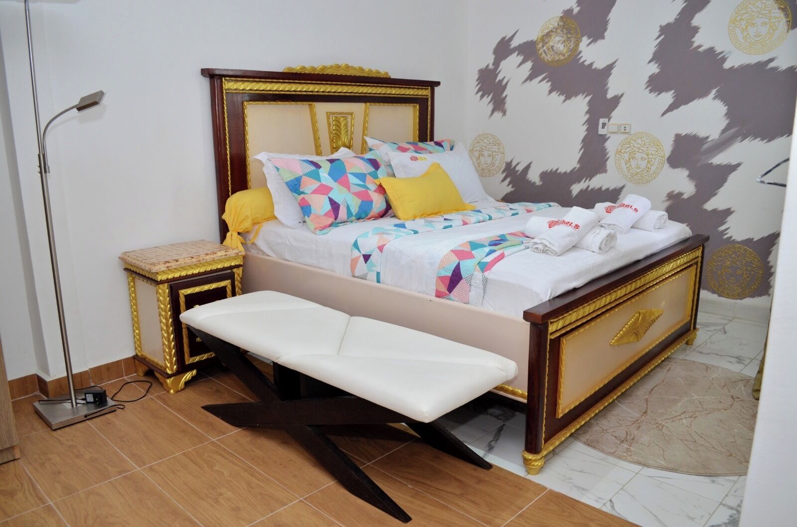 A bed with a bench, a bedside table, and a floor lamp in a room with patterned wallpaper and tiled flooring.