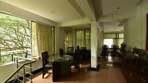 A spacious dining area with a glass table and chairs, large windows with curtains, and a kitchen area with buffet setup. - ANANYA COMFORTSS-OPP RAHEJA RESIDENCY  (Bengaluru)