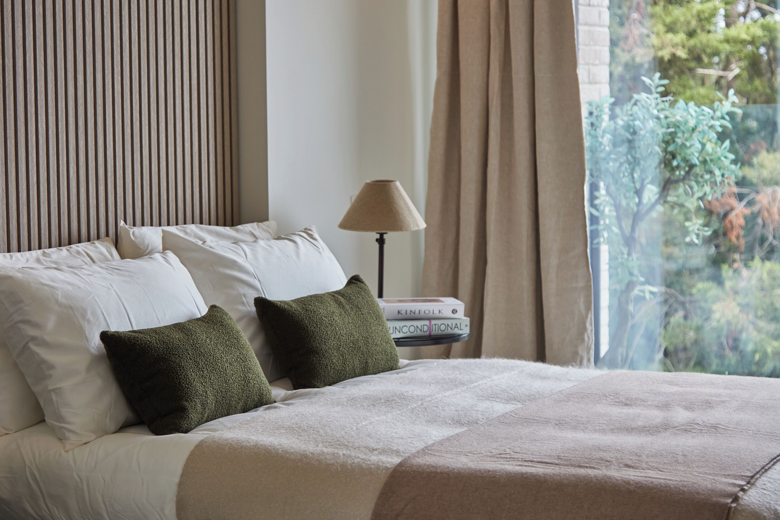 A neatly made bed with white and green pillows, a beige blanket, and a lamp on a bedside table.