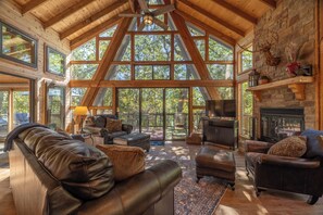A spacious living room with a high ceiling, wooden beams, large windows, a fireplace, and leather furniture. - Meadow Valley Hideout by Innsbrook Vacations! (Innsbrook)