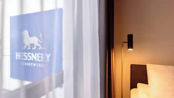 Ein Hotelzimmer mit einem Bett, einem Nachttisch und einer Lampe. Ein Fenster mit dem Schriftzug "Hessney Presidents Club" ist ebenfalls vorhanden.