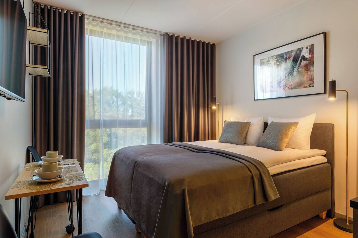 Une chambre d’hôtel moderne avec un grand lit, un bureau avec une lampe et une fenêtre avec des rideaux.