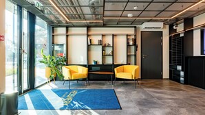 Een moderne lobby met een blauw tapijt, twee gele stoelen, een plant en een boekenplank.