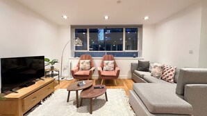 Ein modernes Wohnzimmer mit einem Flachbildfernseher, zwei Sesseln, einem Sofa, einem Couchtisch und einem großen Fenster mit Blick auf die Stadt.