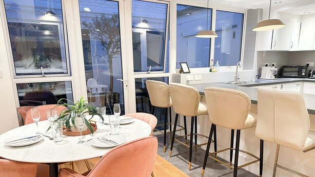 Eine moderne Küche mit Essbereich, mit einem runden Tisch und rosa Stühlen, einer Bar mit Hockern und großen Fenstern mit Blick nach draußen.