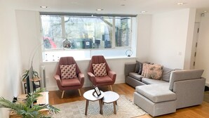 Ein modernes Wohnzimmer mit zwei Sesseln, einem grauen Sofa, einem kleinen Couchtisch und einem großen Fenster mit Blick auf die Straße.