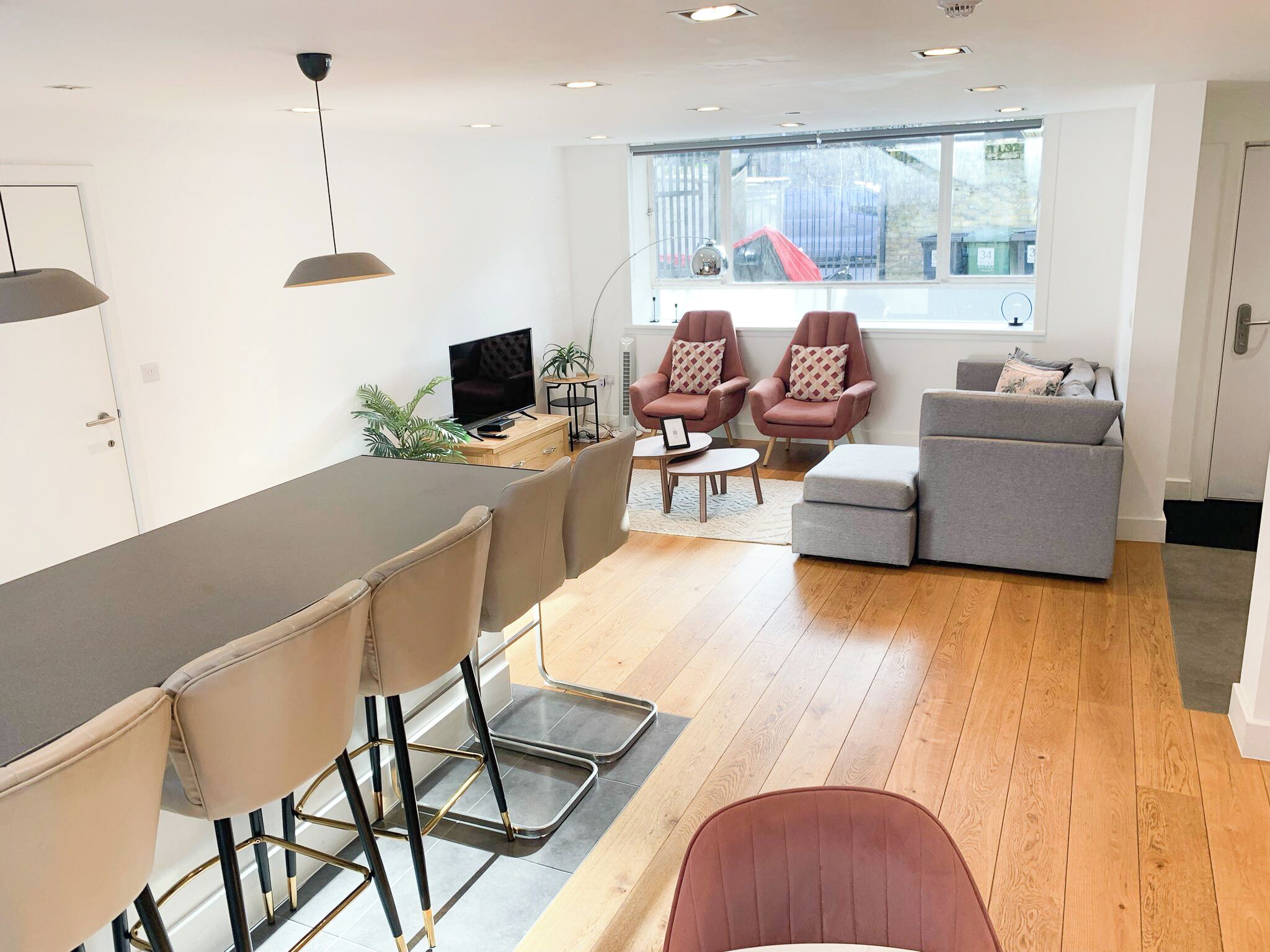 A modern living room with a dining area, a grey sofa, two armchairs, a TV, and a window with a view of the outside.