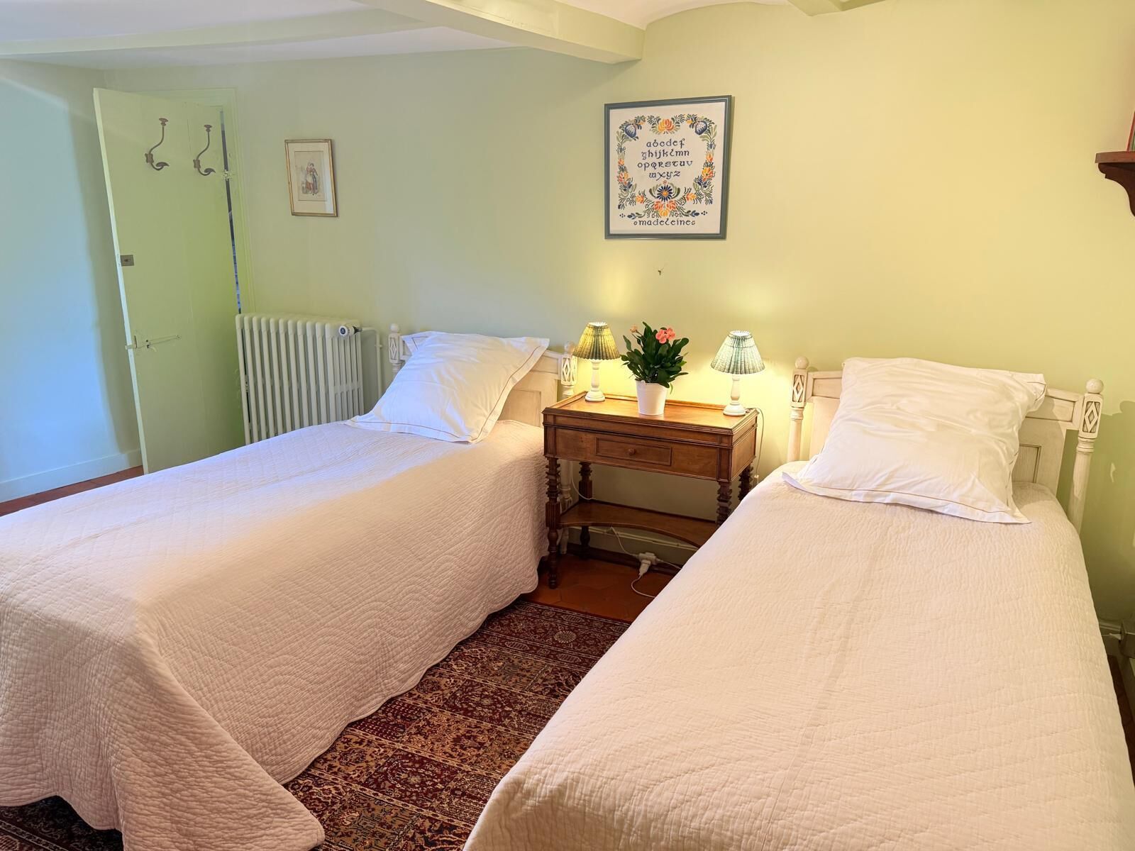 Une chambre avec deux lits simples, un radiateur, une petite table avec des lampes et une affiche illustrant l’alphabet, encadrée, accrochée au mur.