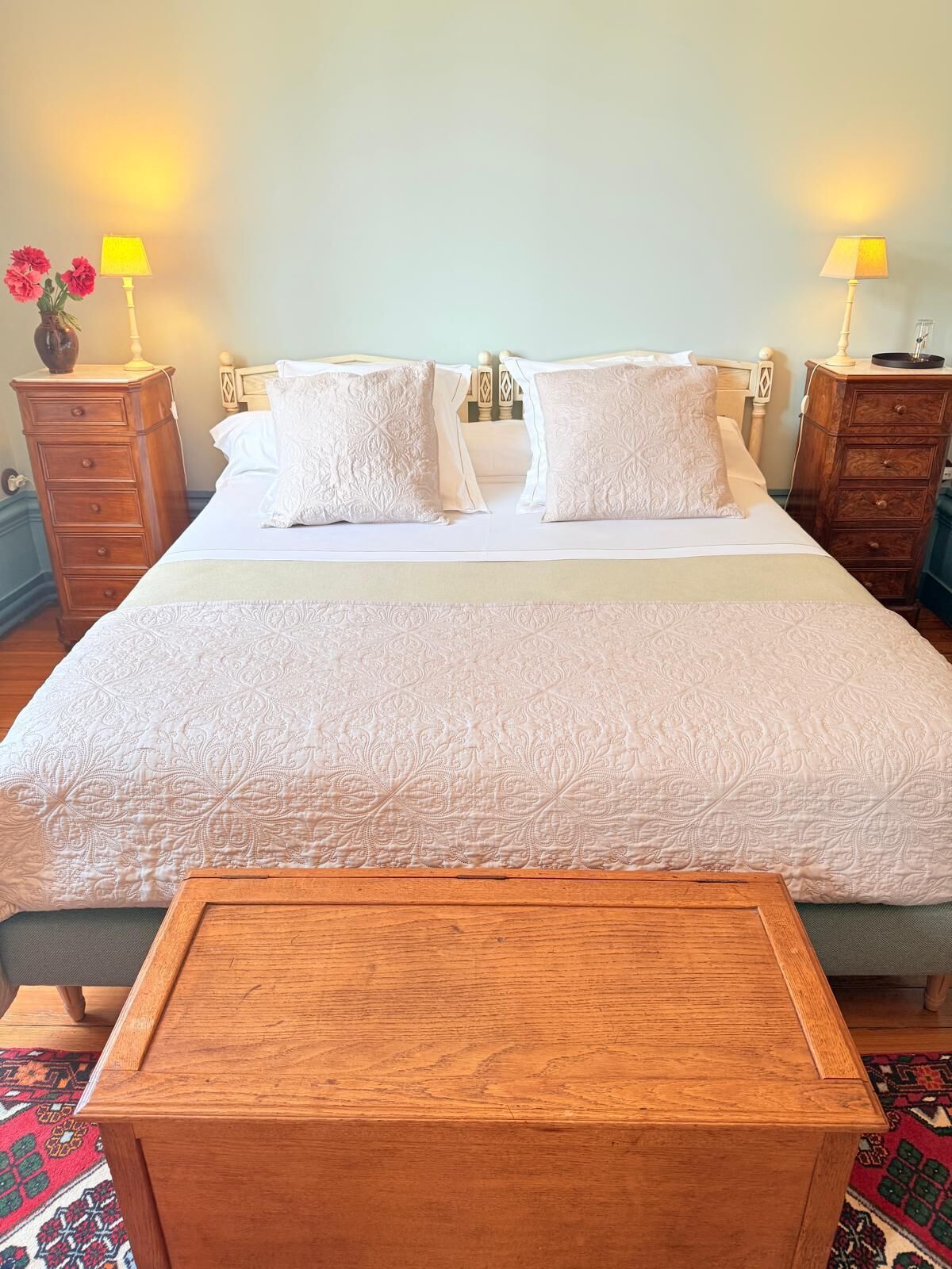 A neatly made bed with a patterned bedspread, two bedside tables with lamps, a wooden nightstand, and a small wooden chest in the foreground.