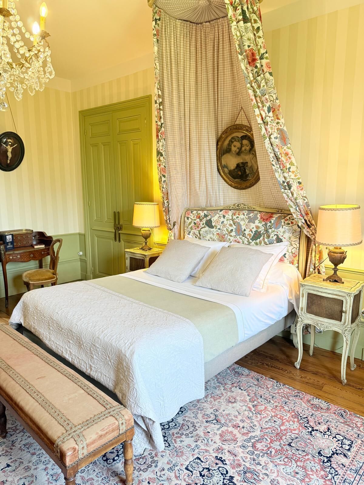 Un dormitorio con cama con dosel, una puerta verde, una mesita con lámpara y una alfombra estampada.