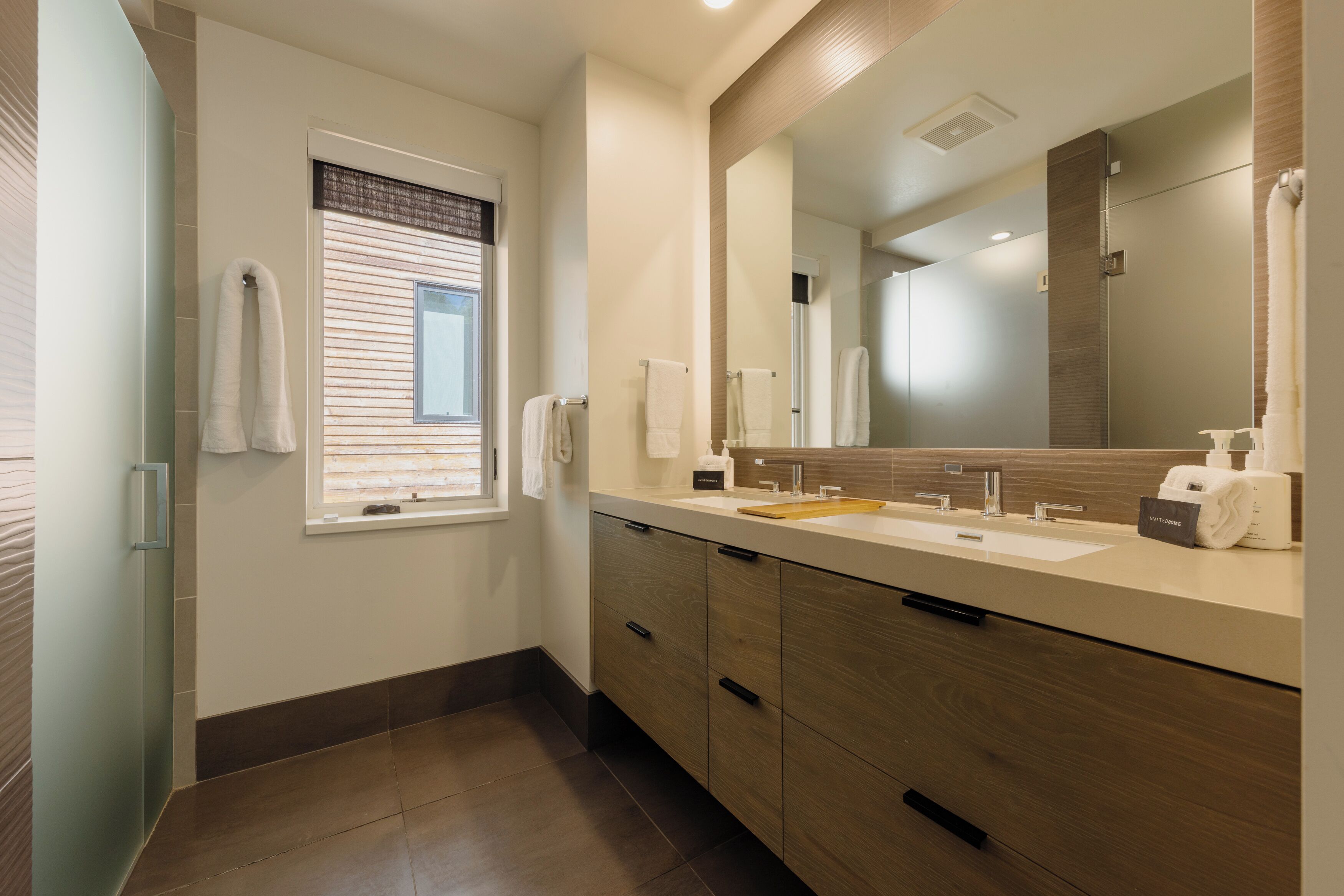 Un baño moderno con un espejo grande, dos lavamanos y una ventana con persianas.
