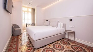A modern hotel room with a large bed, a chair, a small table, and a TV mounted on the wall. - Sweett | Gran Via 204 (Barcelona)
