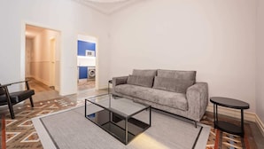 A living room with a grey sofa, a glass coffee table, and a black side table. There is a patterned rug on the floor and a hallway leading to a kitchen with blue cabinets. - Sweett | Gran Via 204 (Barcelona)