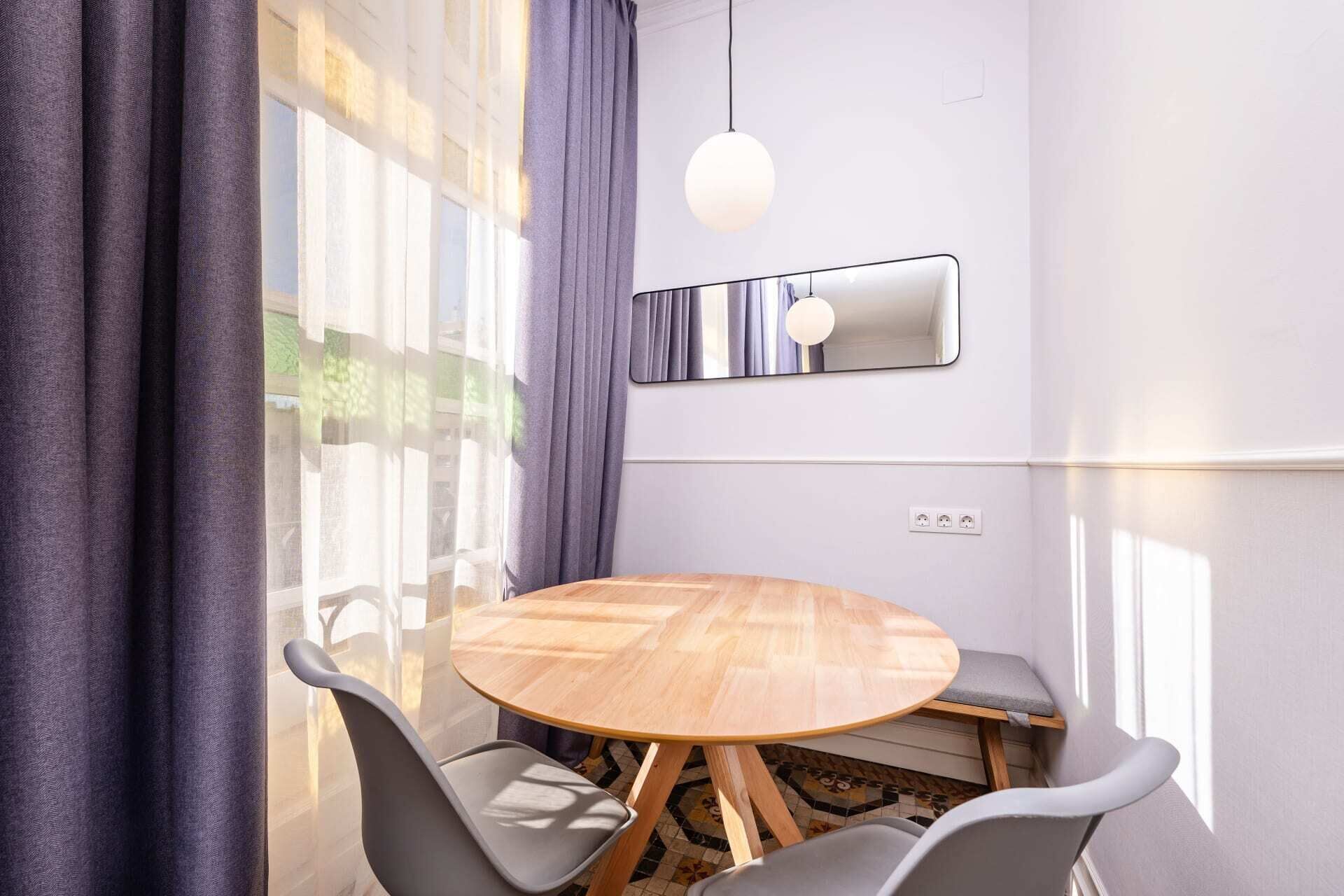 A modern dining area with a round wooden table, grey chairs, a hanging light fixture, and a large window with curtains.