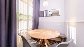 A modern dining area with a round wooden table, grey chairs, a hanging light fixture, and a large window with curtains. - Sweett | Gran Via 302 (Barcelona)