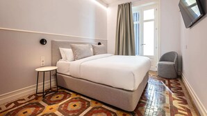 A hotel room with a bed, a small table, a chair, a TV mounted on the wall, and a patterned floor. - Sweett | Gran Via 302 (Barcelona)