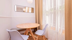 A modern dining area with a round wooden table, two white chairs, a hanging light fixture, and a large window with sheer curtains. - Sweett | Gran Via 104 (Barcelona)