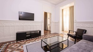 A modern living room with a flat-screen TV mounted on the wall, a glass coffee table, a black entertainment unit, a grey sofa, and a patterned carpet. - Sweett | Gran Via 202 (Barcelona)