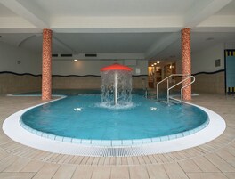 Ein Innenpool mit einem zentralen Wasserspiel, umgeben von gefliesten Böden und Säulen mit Mustern.