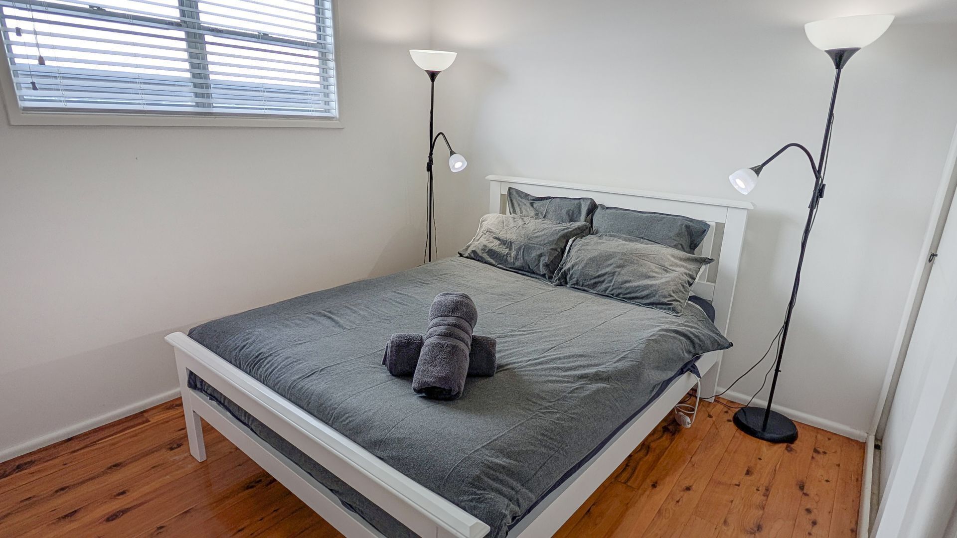 A bedroom with a bed, two bedside lamps, a window with blinds, and a wooden floor.