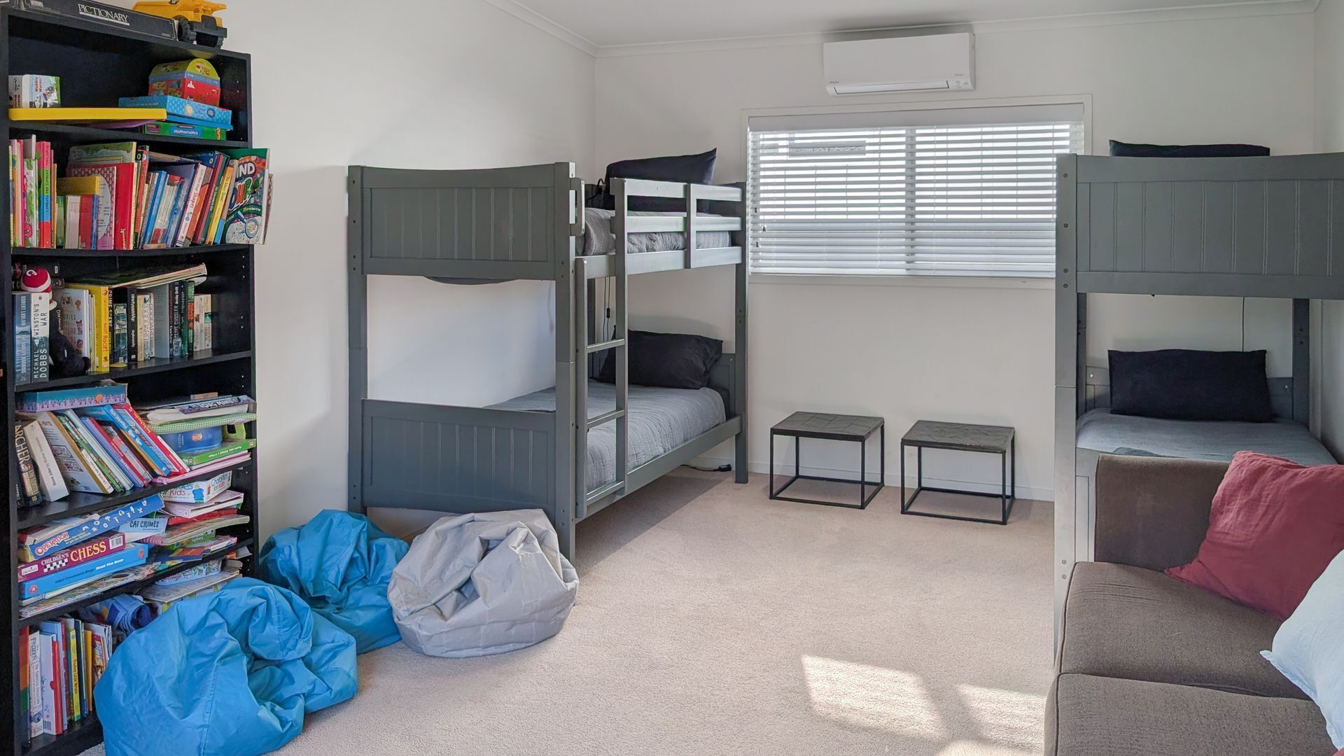 A bunk bed room with a bookshelf, two small tables, and a window with blinds.
