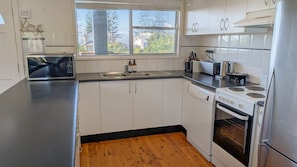 Cozinha com armários brancos, geladeira de aço inoxidável, fogão, lava-louças e janela com vista para casas e árvores.