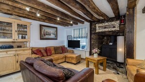 Een gezellige woonkamer met zichtbare houten balken, een open haard, een televisie en een houten kast met glazen deuren.