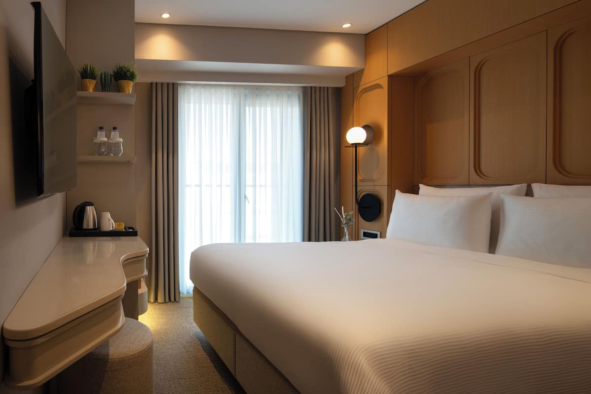 Quarto de hotel com uma cama grande, mesa de cabeceira com chaleira, televisão de tela plana e janela com cortinas.