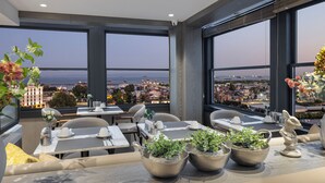 Un área de comedor en la azotea con mesas dispuestas para dos, ventanas amplias que ofrecen vistas a la ciudad y plantas en macetas sobre las mesas.