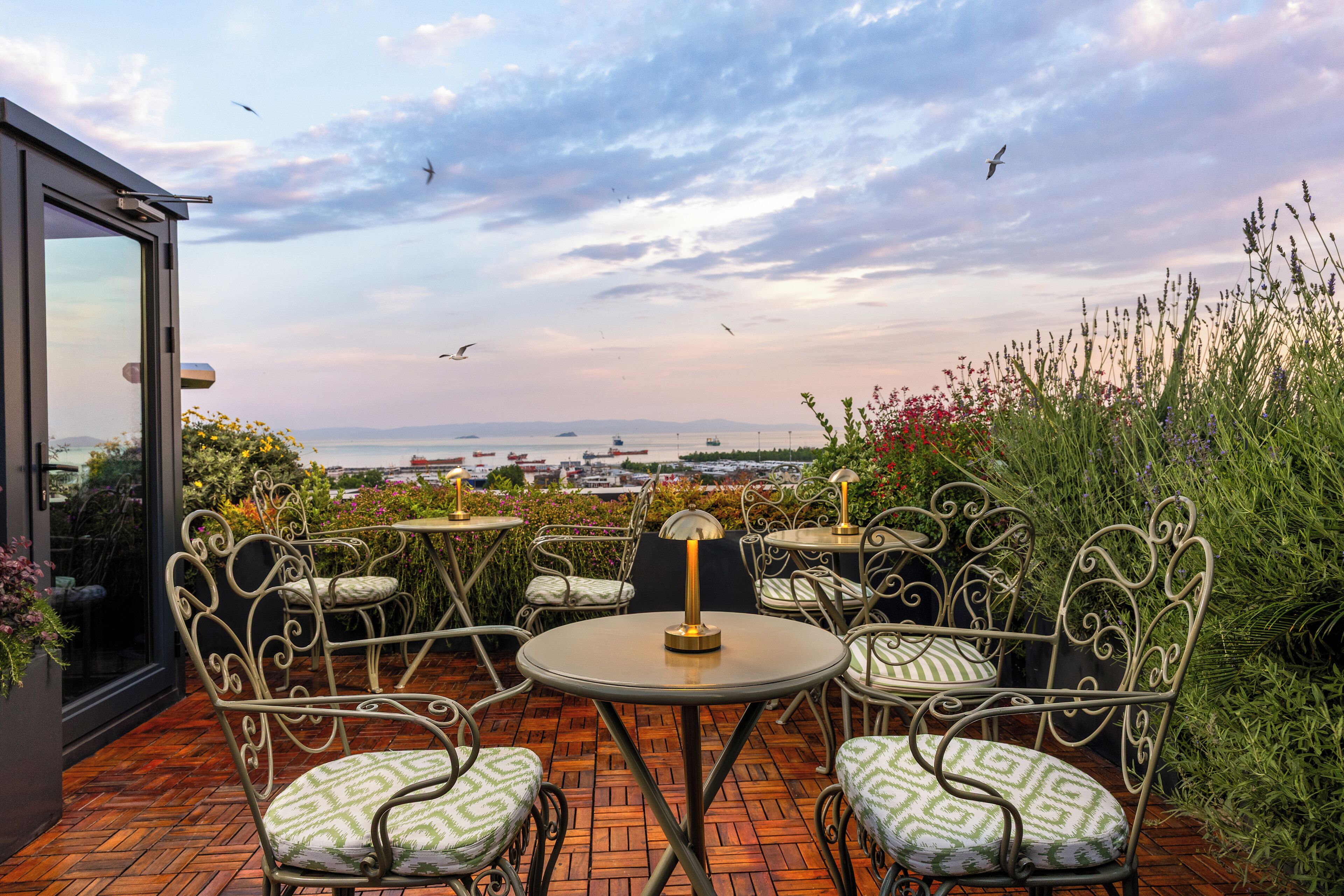 An outdoor patio with metal chairs and tables, a view of the sea, and a glass door leading to the interior.