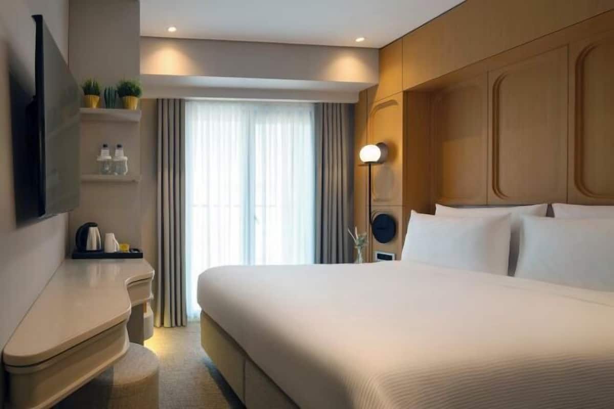 Quarto de hotel com uma cama grande, mesa de cabeceira, luminária, televisão na parede e uma janela com cortinas.