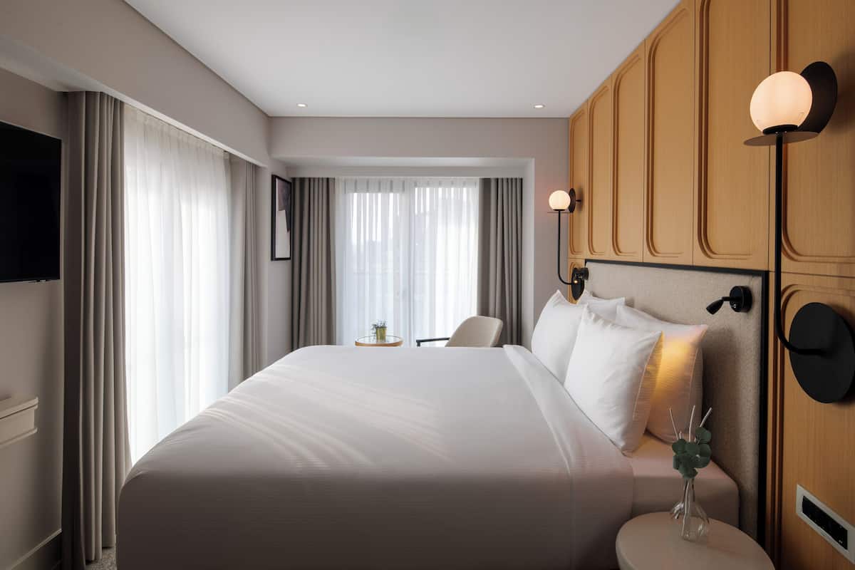 Quarto de hotel com uma cama grande, uma mesinha de cabeceira com abajur, uma TV na parede e uma janela com cortinas.