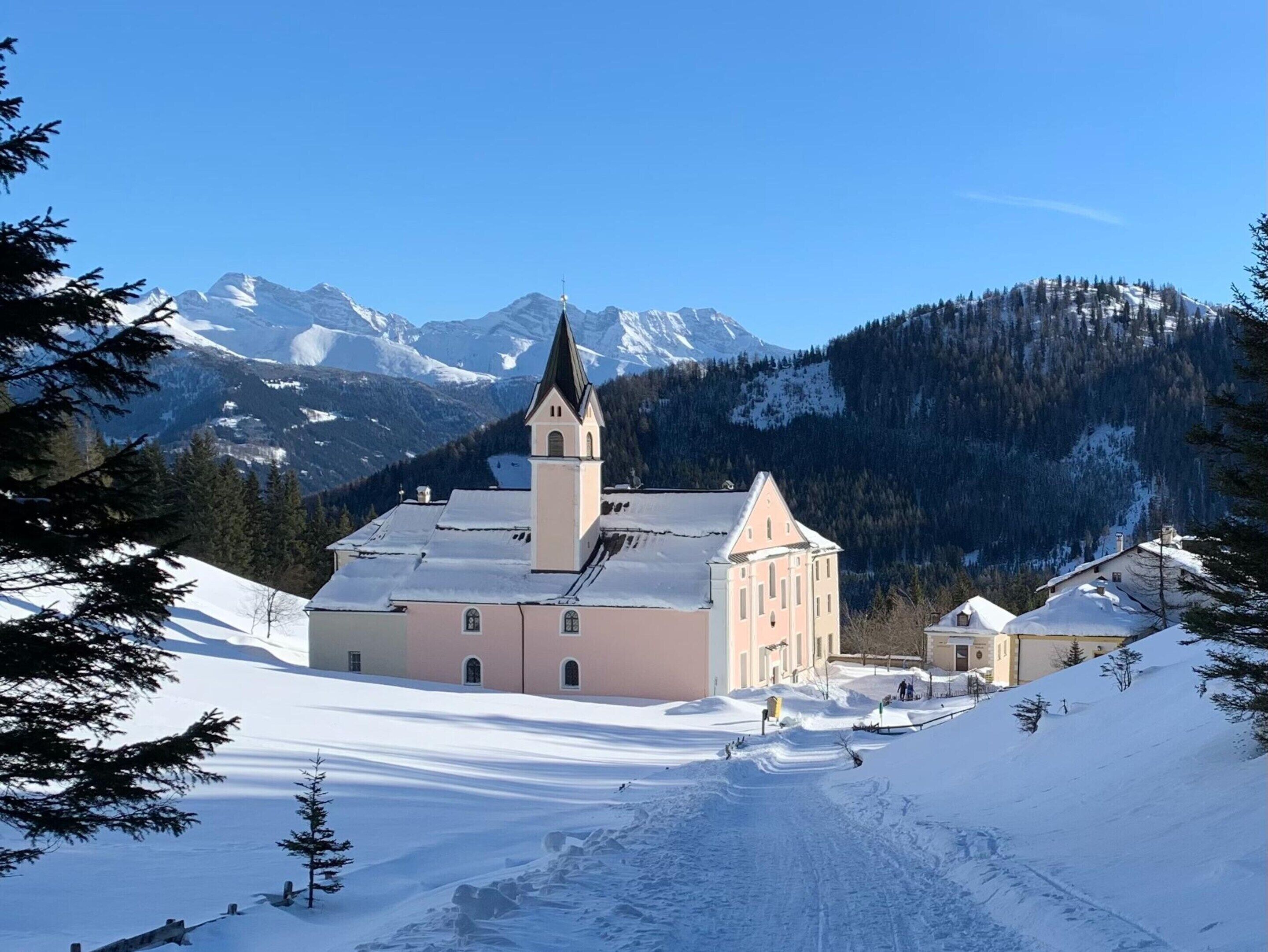 A church with a tall steeple and a bell tower, surrounded by snow-covered mountains and trees.