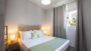 Un lit bien fait, agrémenté d’un plaid vert, une lampe de chevet et une fenêtre avec des rideaux.