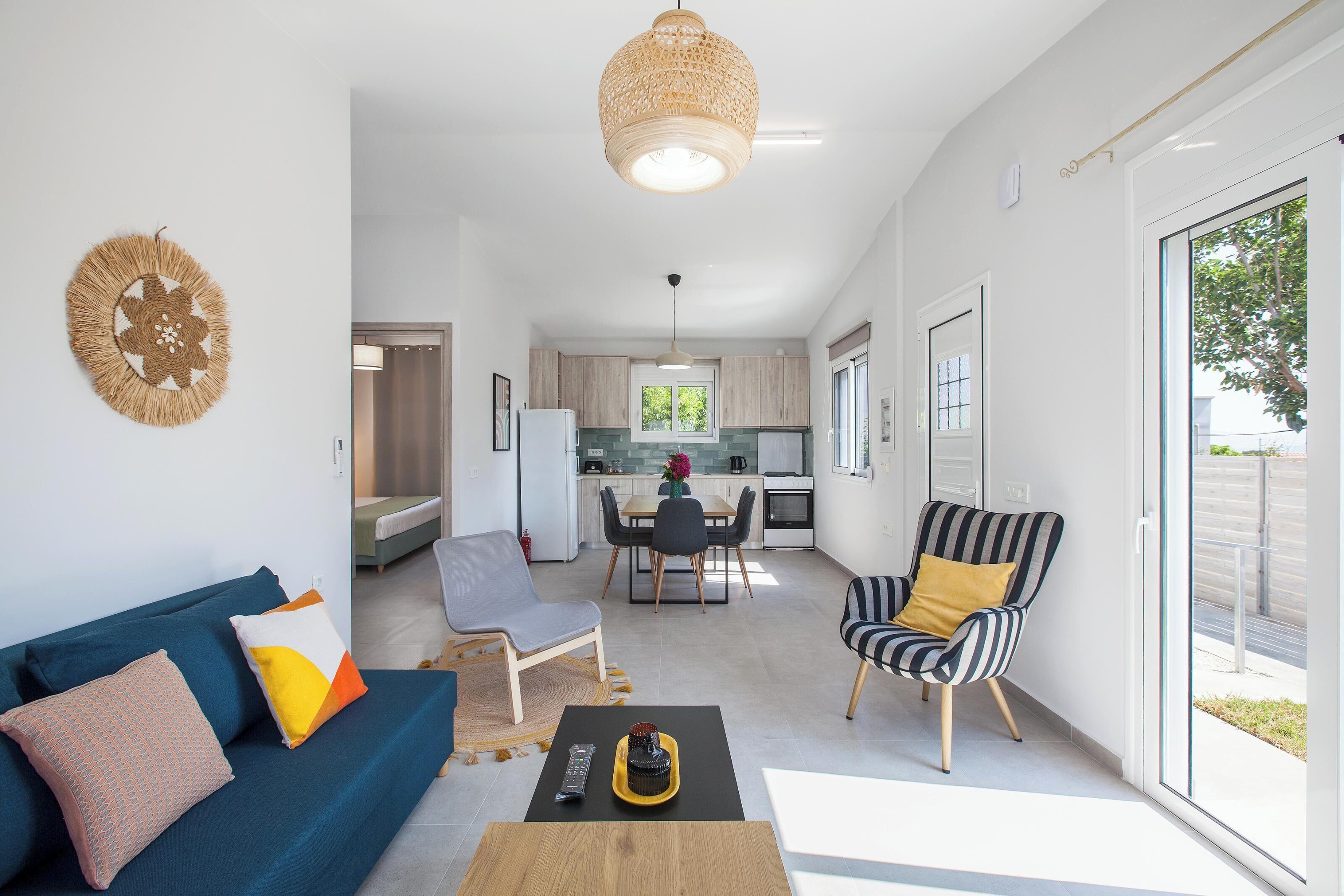 Una sala moderna con un sofá, una mesa de centro y un sillón a rayas. Se ve una cocina al fondo con una mesa de comedor y sillas.