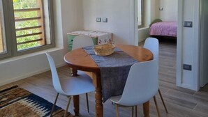 A dining area with a wooden table, white chairs, and a woven basket. There is a window with wooden blinds, a rug, and a bed visible in the background.