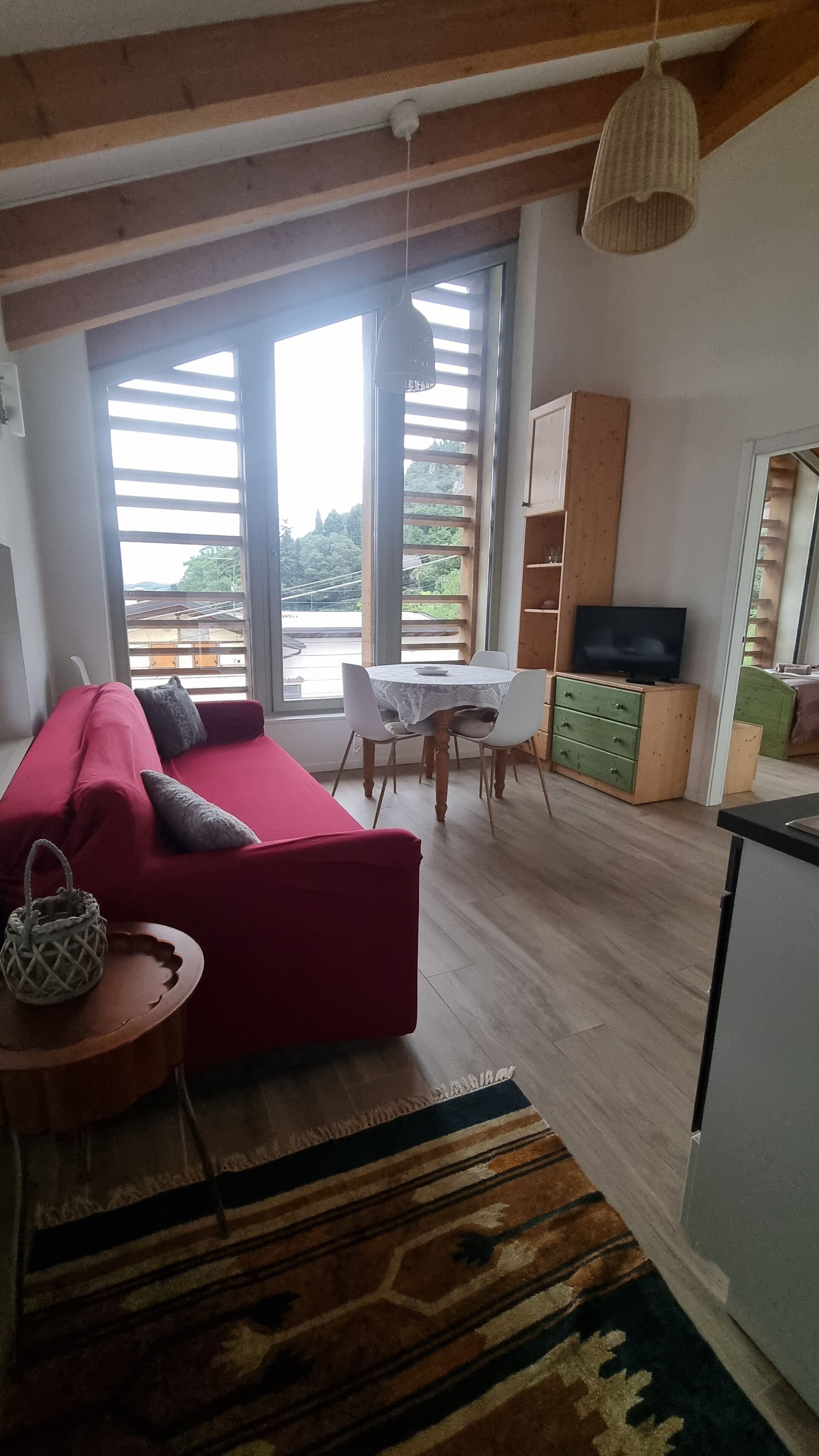 A living room with a red sofa, a small round table, and a TV stand. There is a large window with a view of greenery outside.