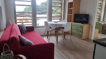 A living room with a red sofa, a small round table, and a TV stand. There is a large window with a view of greenery outside.