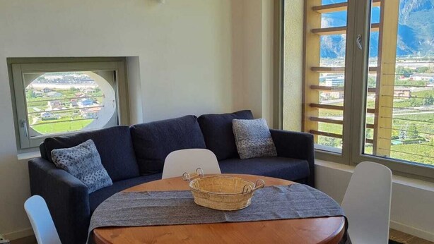 A cozy room with a dark blue sofa, white chairs, a wooden table, and a view of a mountainous landscape through large windows.