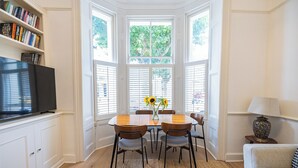 A dining area with a bay window, a table with chairs, a vase with sunflowers, and a television on a stand. - Charming 3-Bed Haven in Little Venice with a Private Outdoor Oasis (London)