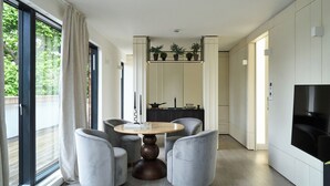 A modern dining area with a round wooden table surrounded by four grey upholstered chairs, a black dining table in the background, and a TV mounted on the wall. - Loftus Road (London)
