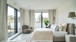 A modern bedroom with a large bed, a sofa, a chair, and a view of the city through large windows. - Loftus Road (London)