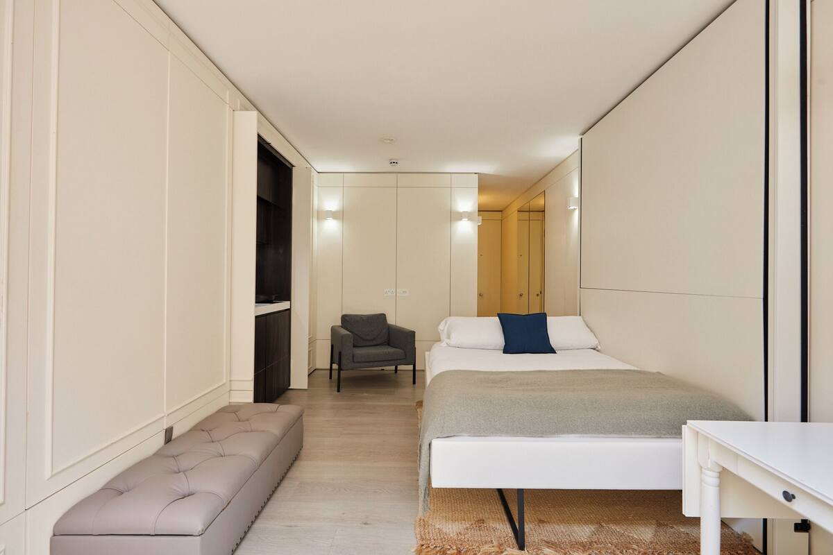 A narrow hotel room with a bed, a bench, a small table, and a chair.