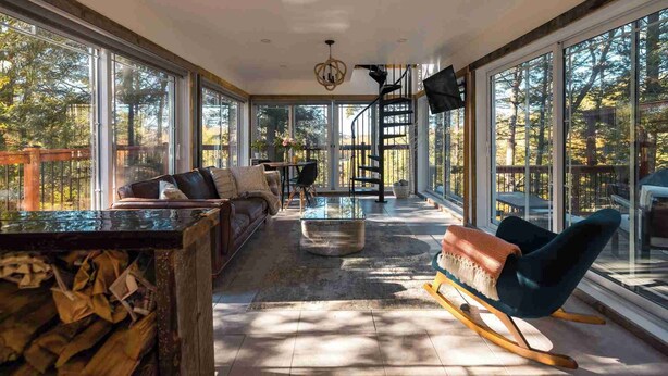 Una habitación amplia con ventanas grandes, una escalera curva, un sofá, un sillón mecedora y una mesa de centro de vidrio.