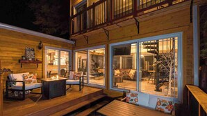 Terraza de madera con mobiliario exterior, amplios ventanales correderas y un espacio interior visible con chimenea.