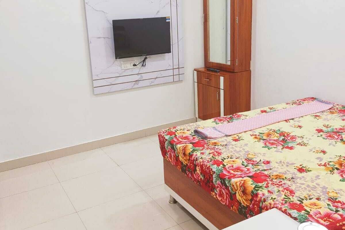 Una camera da letto con un letto, una TV montata su una parete in marmo, un armadio in legno, un comodino con un bollitore e uno specchio.