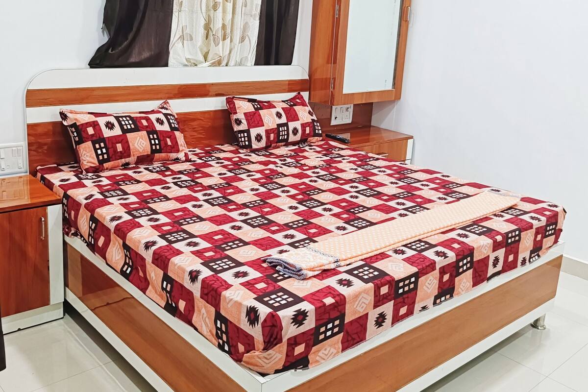 Un letto rifatto con cura, con copriletto a fantasia, una testata in legno e un comodino con una lampada.